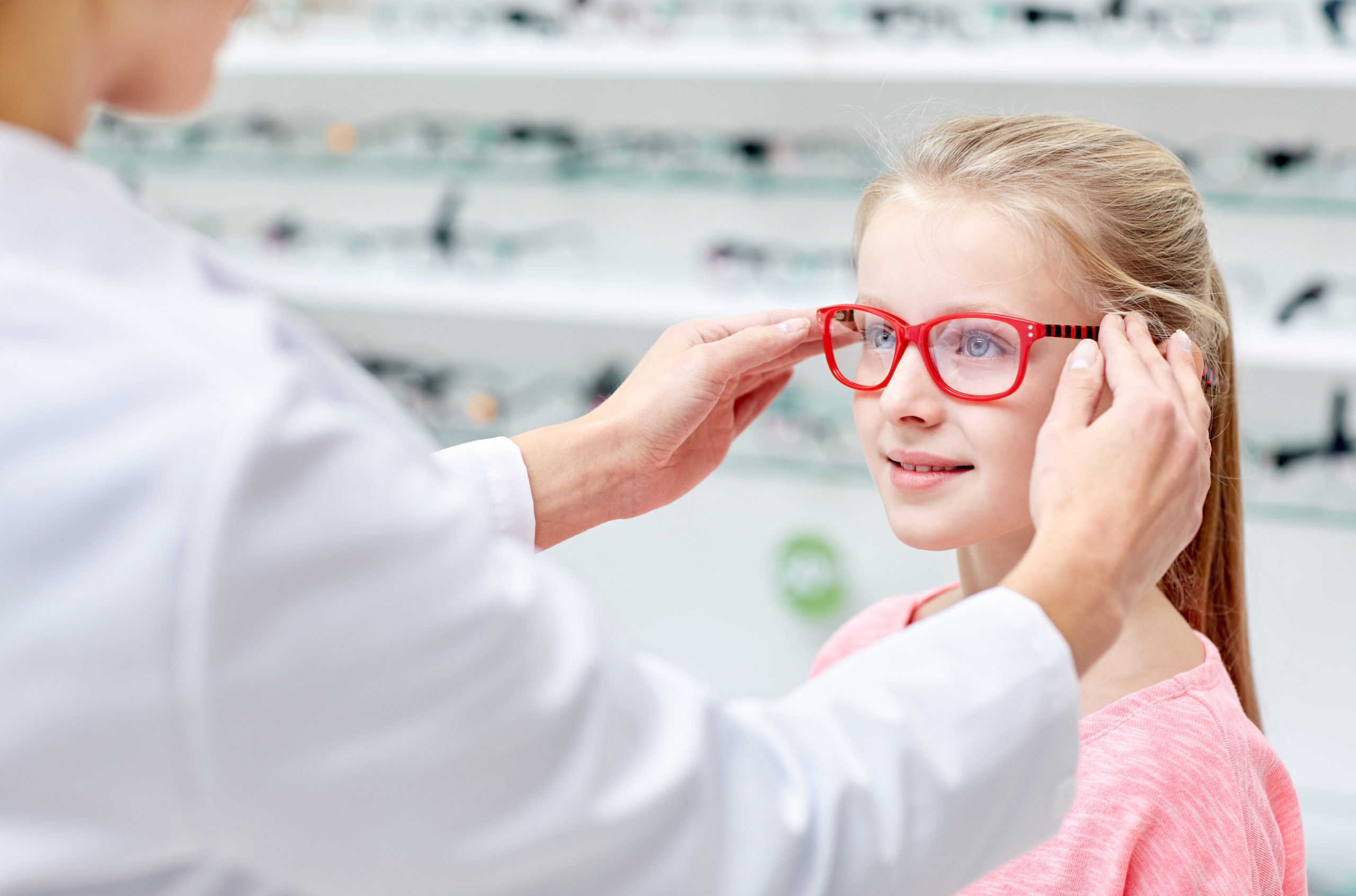 health care, people, eyesight and vision concept - optician putting glasses to little girl eyes at optics store