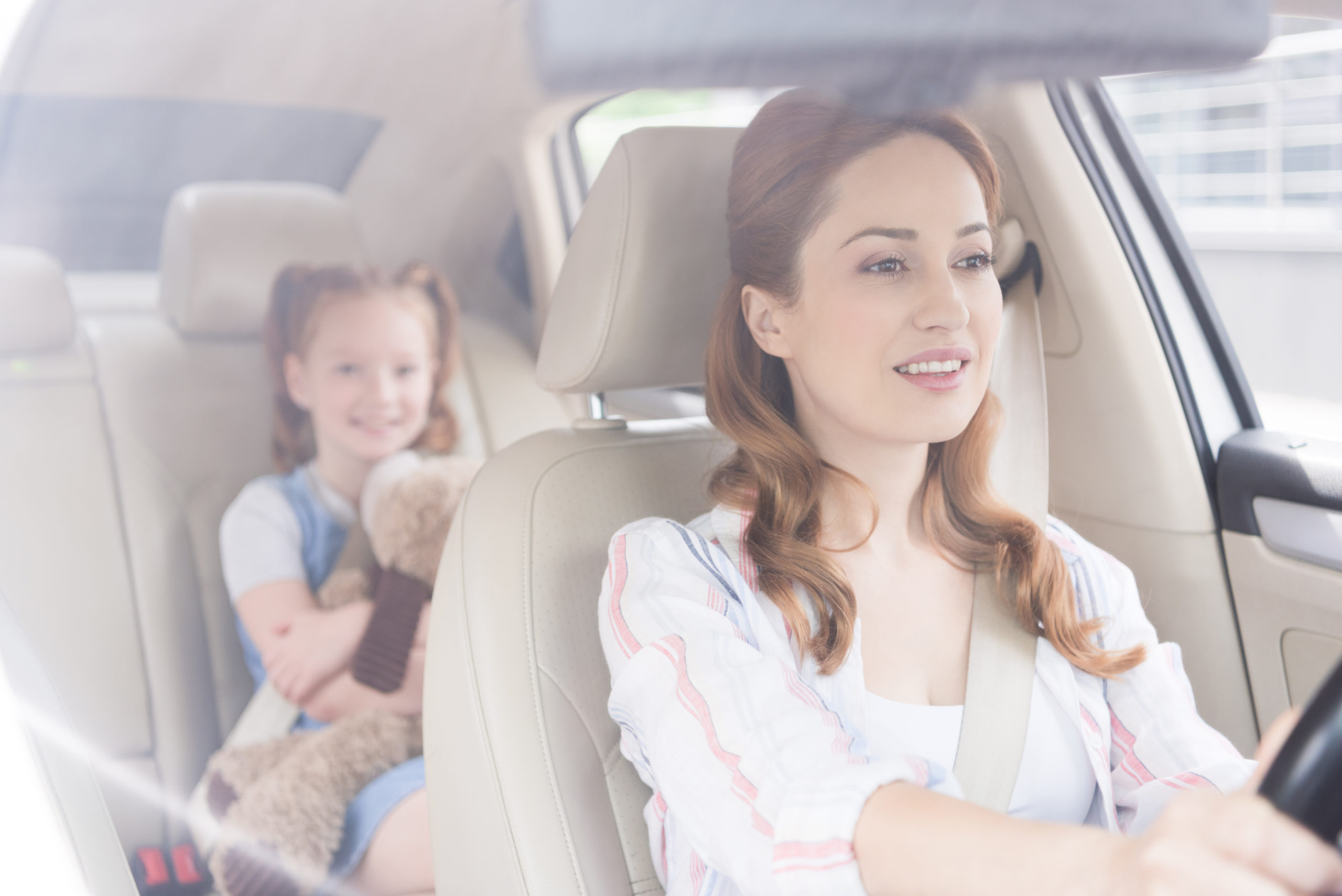 selective focus of smiling mother driving car with daughter on passengers seat behind