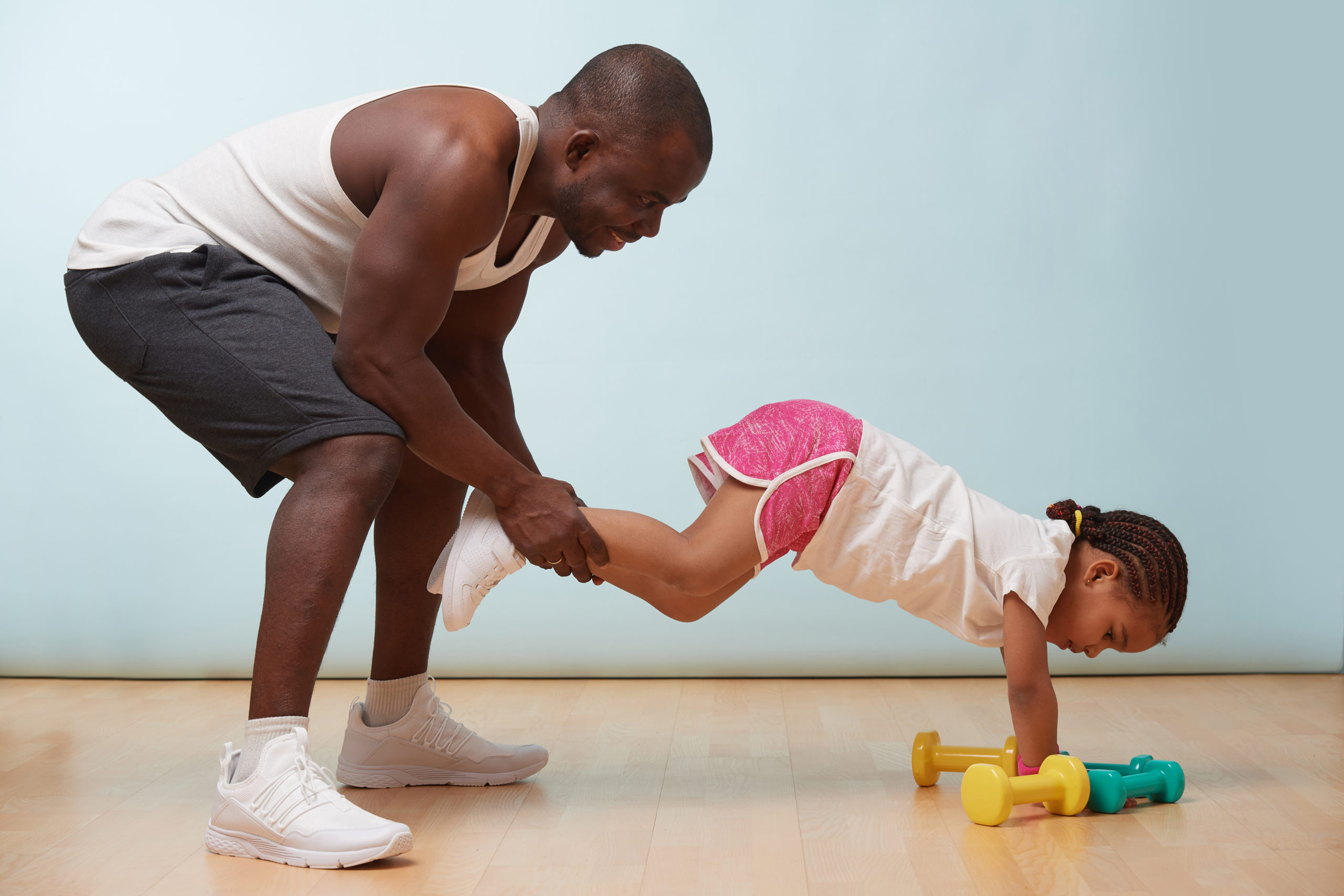 Handsome black young father is helping his cute little daughter to exercise at home. He is holding her legs so she would try advanced elevated push ups.