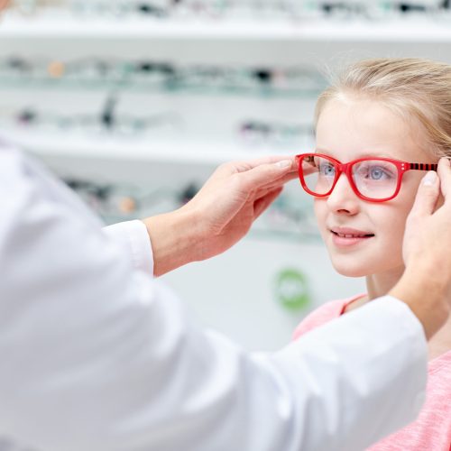 health care, people, eyesight and vision concept - optician putting glasses to little girl eyes at optics store